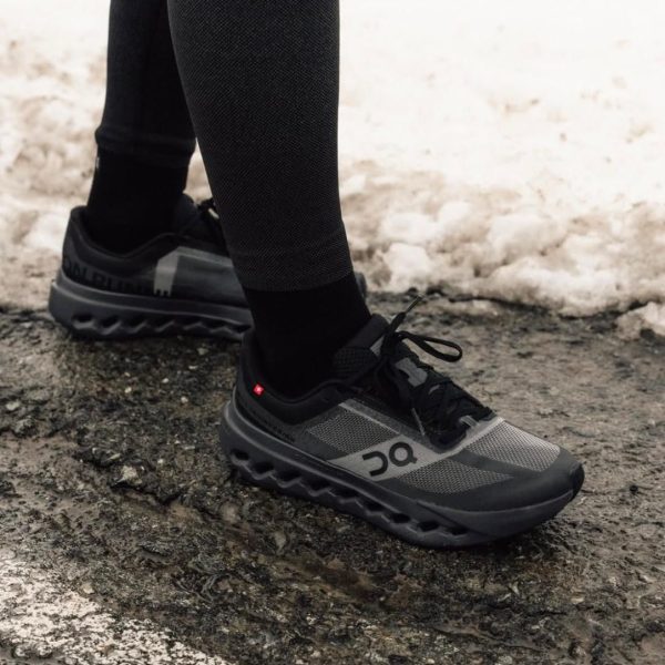Close-up of a person wearing gray and black running shoes and black leggings stepping on a wet pavement with snow in the background.