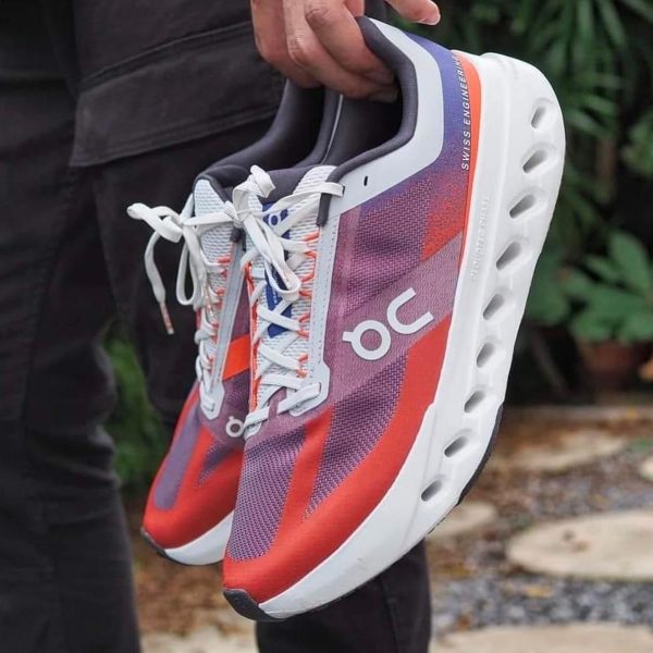 Pair of multicolor running shoes (pink, purple, orange) with white laces held by the heel in front of a garden backdrop.