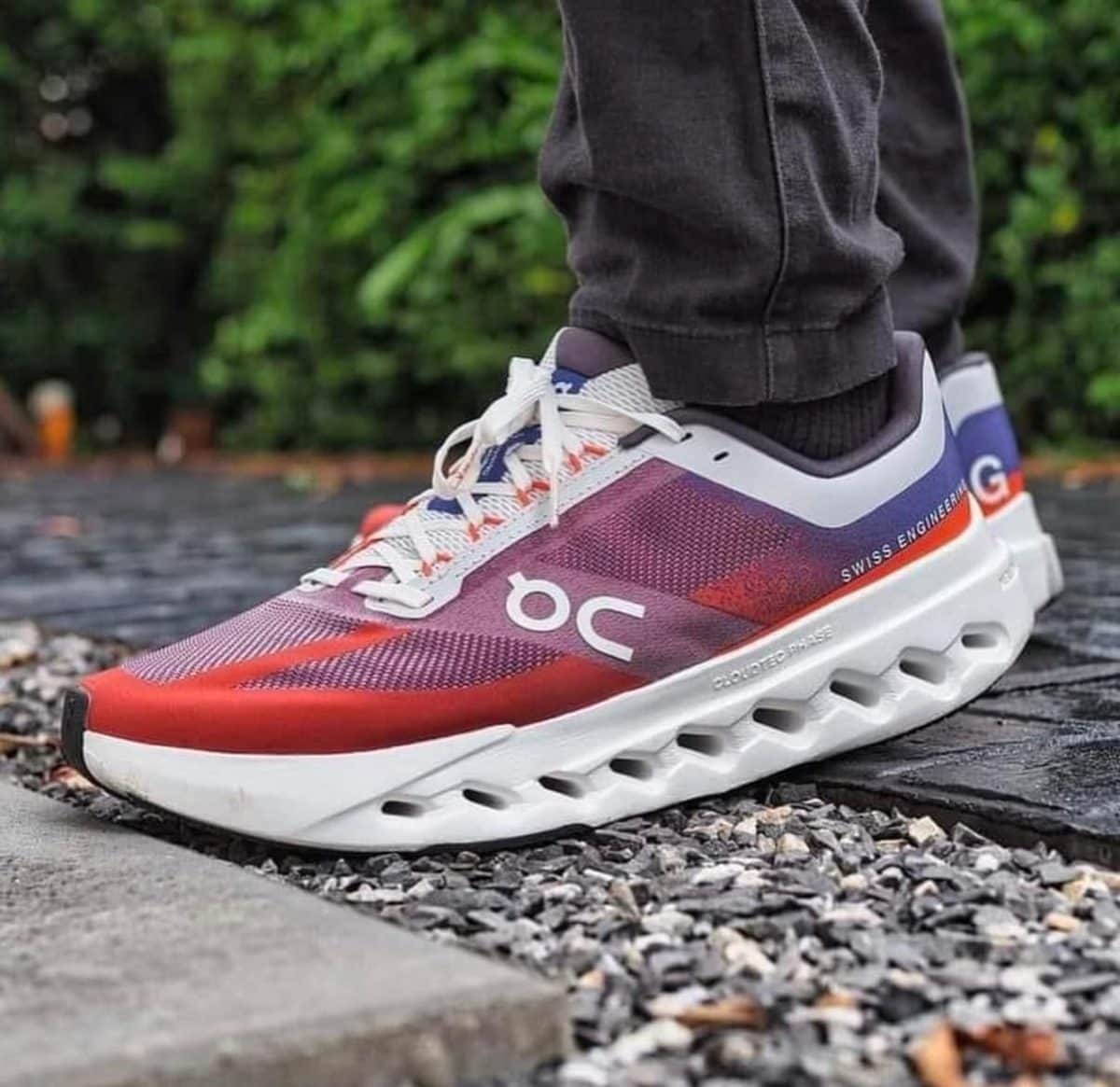 Close-up side view of a purple-pink gradient running shoe with white laces and a chunky white sole on a gravel path, worn by a person in dark pants; brand logo visible.