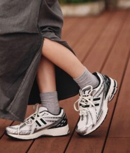 WhatsApp_Image_2025-02-13_at_14.22.46 | UAE SPORT STORE Close-up of a person’s legs and feet wearing white, black, and gray athletic sneakers with gray socks, resting on a wooden deck.