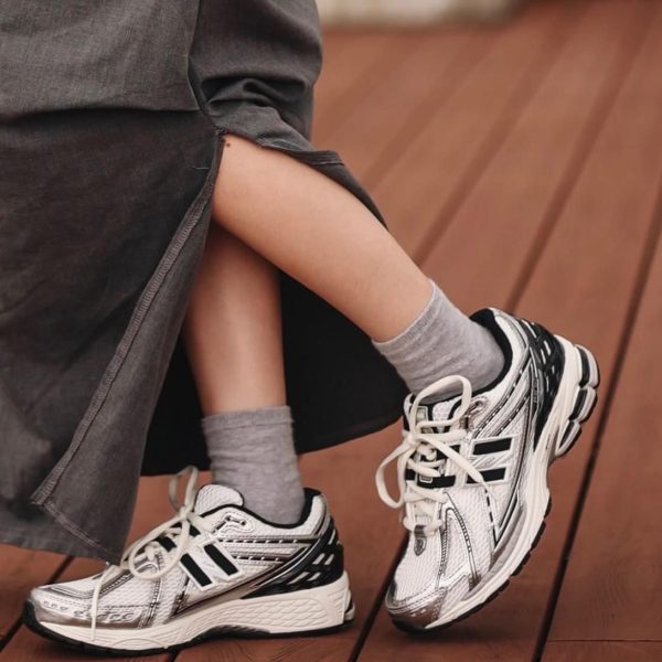 Close-up of a person’s legs and feet wearing white, black, and gray athletic sneakers with gray socks, resting on a wooden deck.