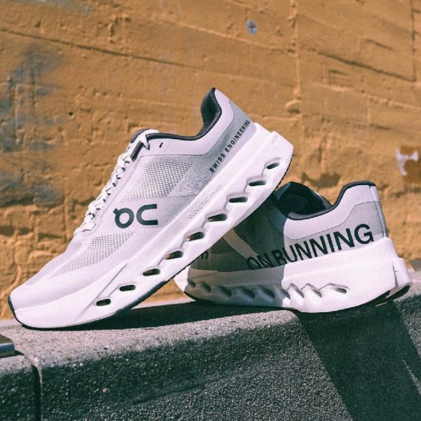 Pair of white On Running sneakers with black accents perched on a rooftop edge against a yellow brick wall, showing the Swiss engineering branding on the side.