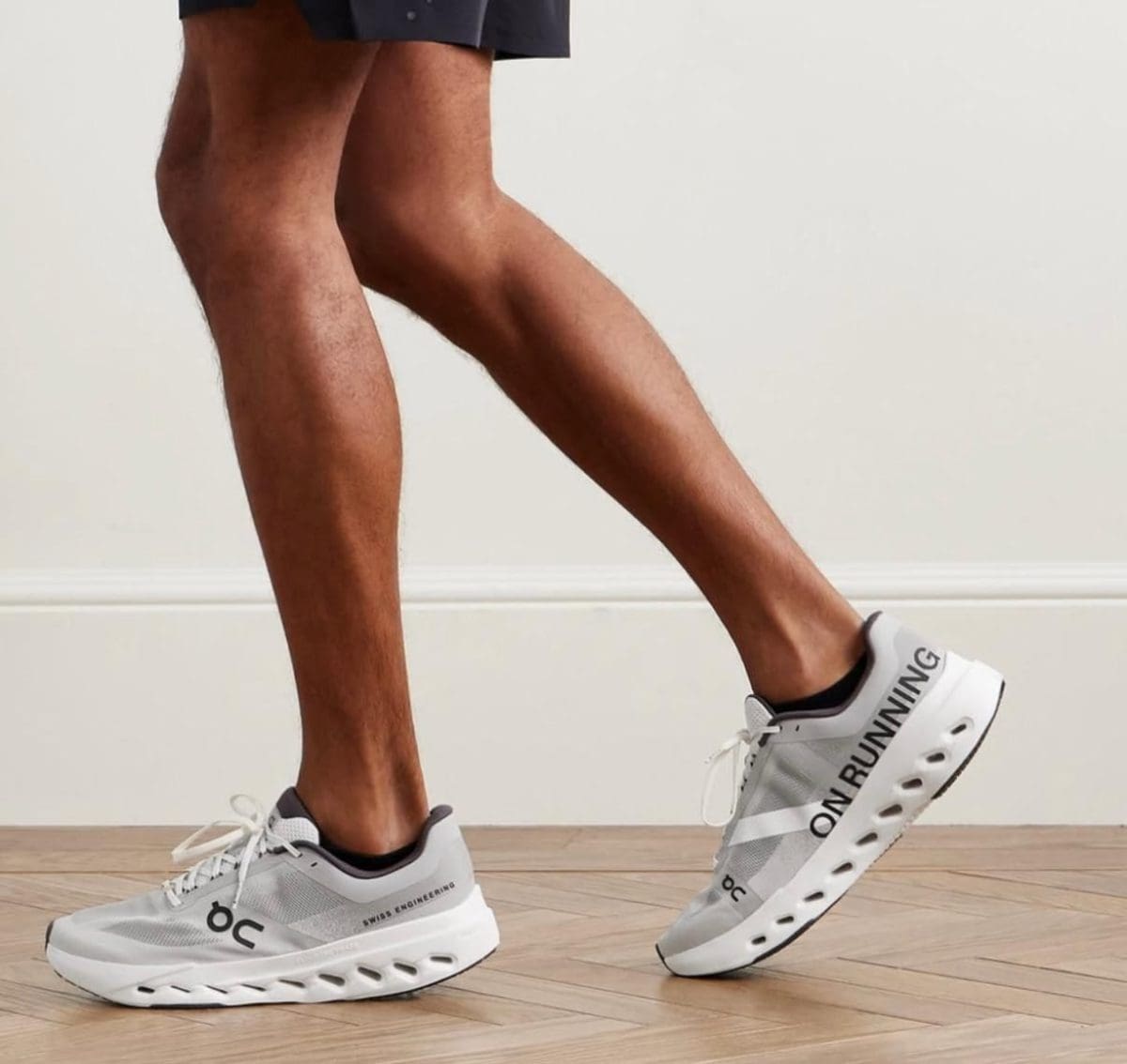 Person wearing white and gray On Running sneakers mid-stride on a wooden floor indoors.