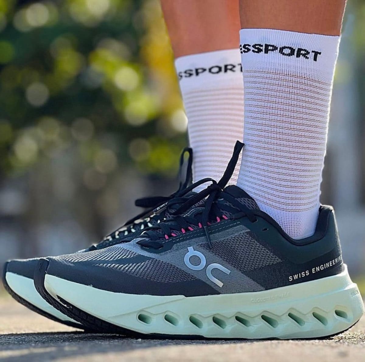 Close-up of a dark gray athletic running shoe with a pale mint sole, laces tied, worn with white socks showing 'SSPORT' text.