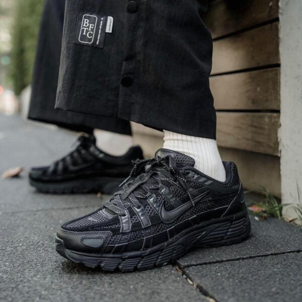 Black Nike running shoes with textured panels, white crew socks, and dark cuffed pants on a sidewalk edge.