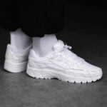 White athletic sneakers worn with white socks, standing on a gray surface in a monochrome shot.