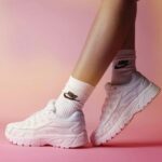 Close-up of white Nike running shoes worn with white socks, standing on a pink surface.