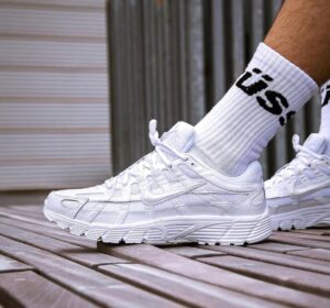White Nike running shoe on a wooden deck, worn with a white crew sock with black lettering visible at the ankle.