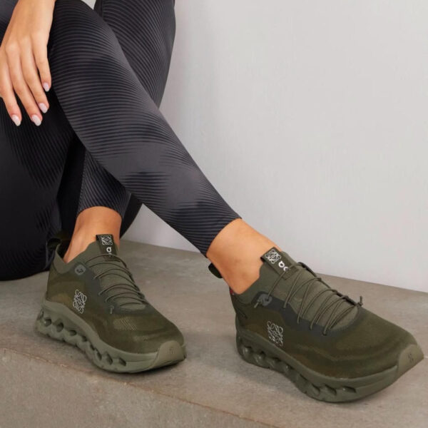 Person seated on a concrete step wearing olive-green athletic sneakers with black patterned leggings. The shoes have a unique chunky sole and laces visible.