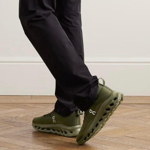 Person wearing olive green On running shoes stepping forward on a wooden floor.