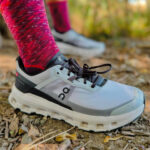 Close-up of a gray On running shoe on a rocky trail, paired with bright pink knit socks; autumn leaves on the ground behind.