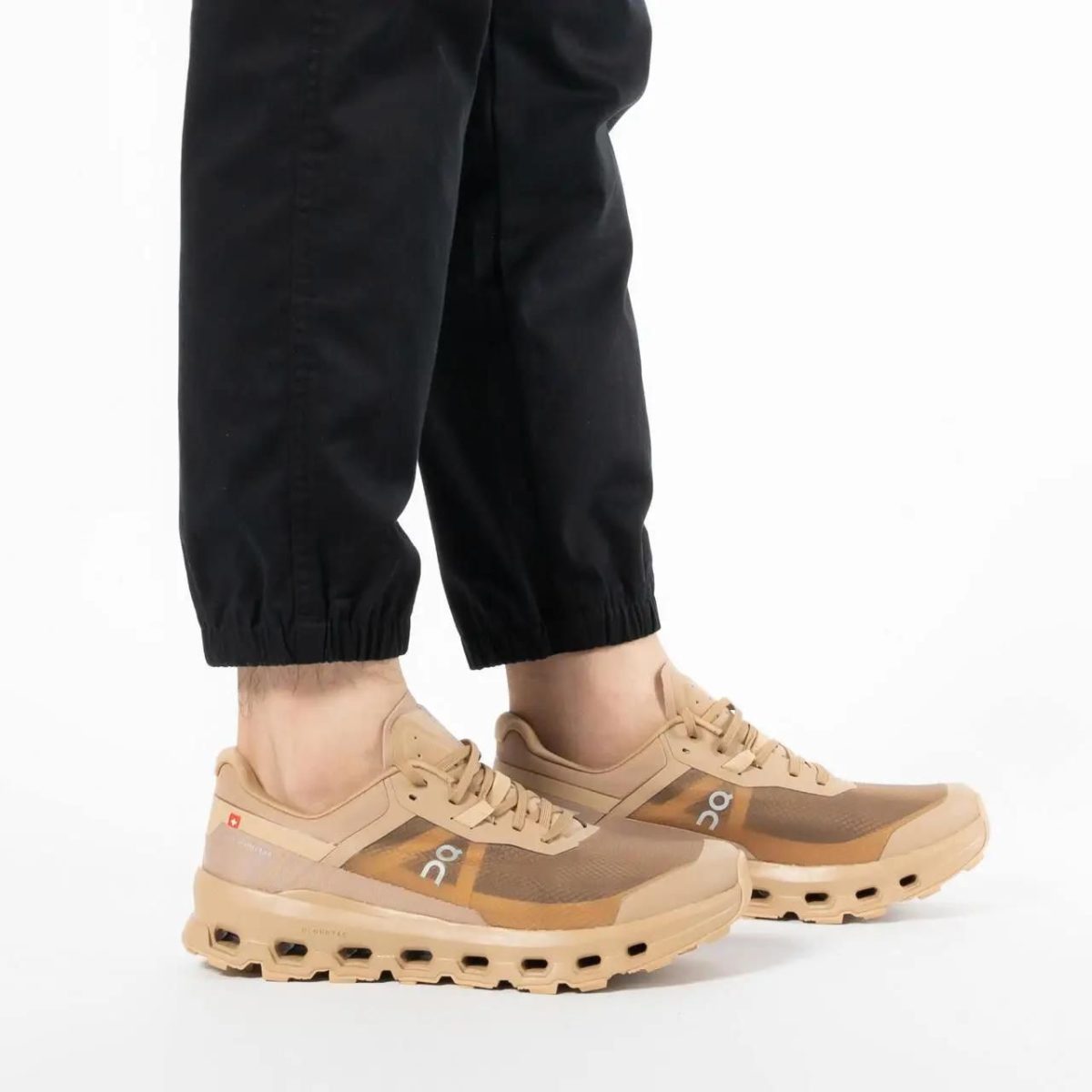 Beige athletic sneakers with a chunky sole worn with black jogger pants on a white background.
