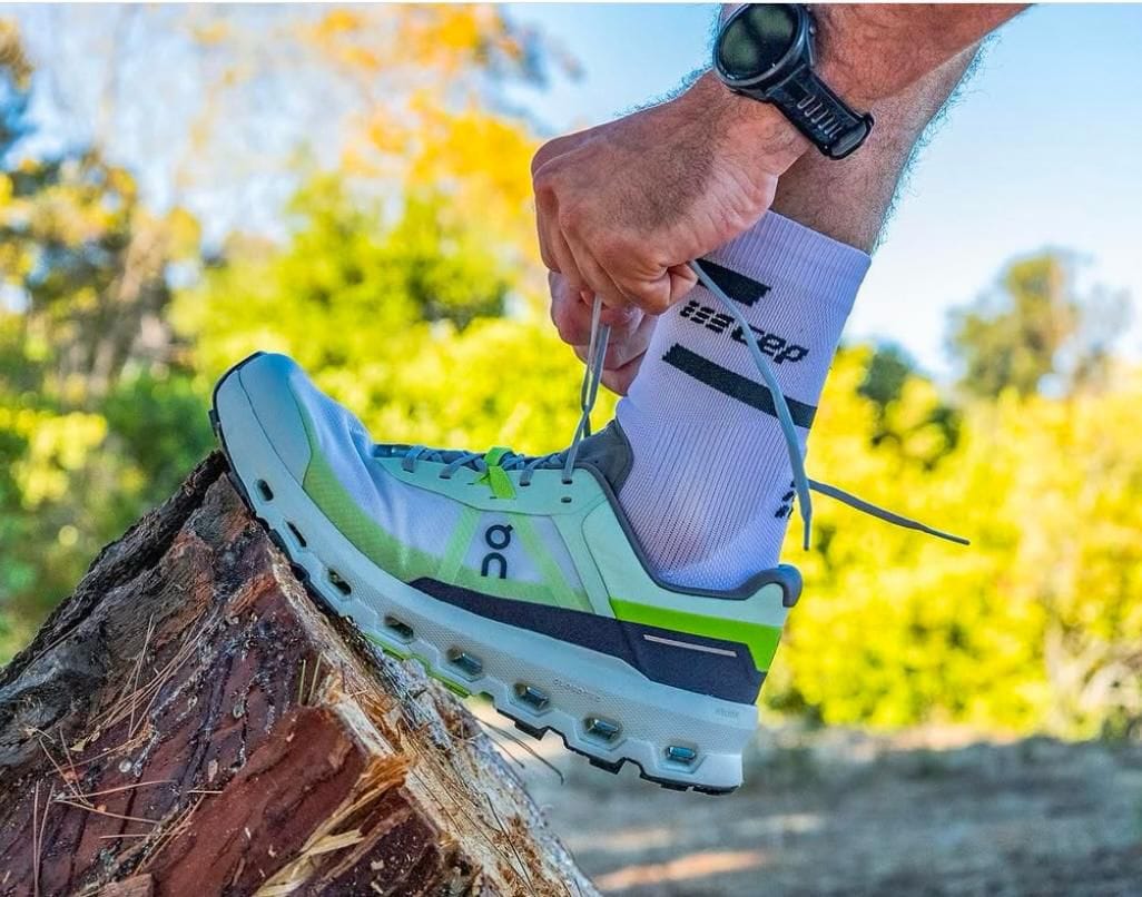 WhatsApp_Image_2025-02-22_at_11.32.46 | UAE SPORT STORE Person tying the laces of a light blue-green running shoe on a tree stump outdoors, wearing a white sock and a sports watch.