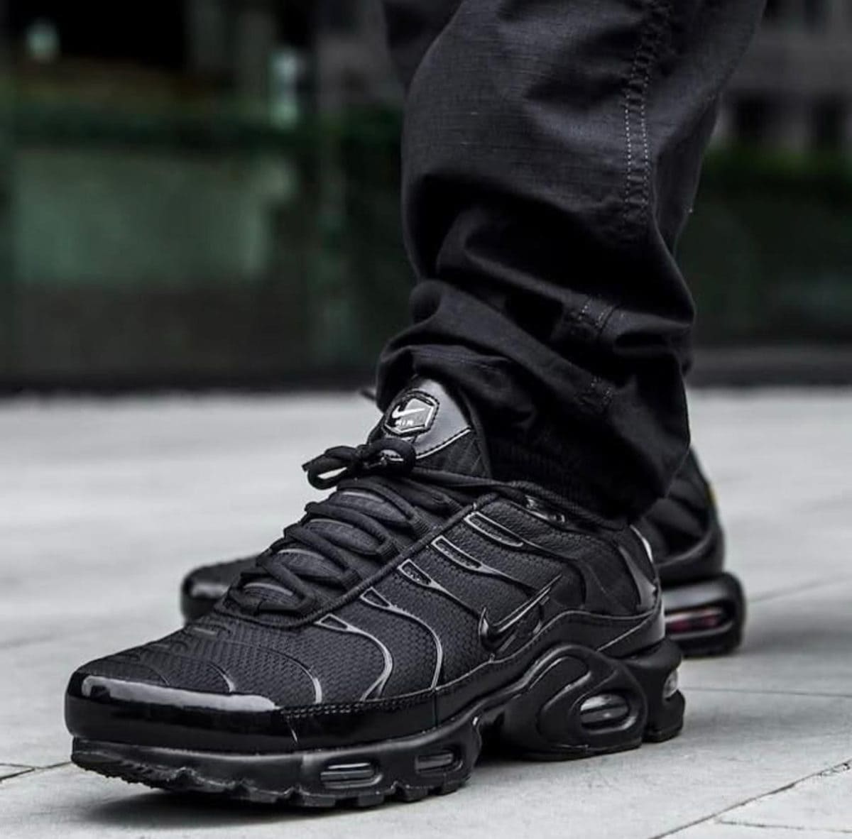 Close-up of a person wearing black Nike sneakers with a visible air midsole, standing on a light pavement surface.