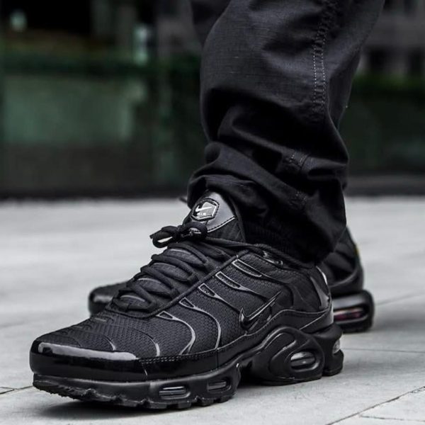Close-up of a person wearing black Nike sneakers with a visible air midsole, standing on a light pavement surface.