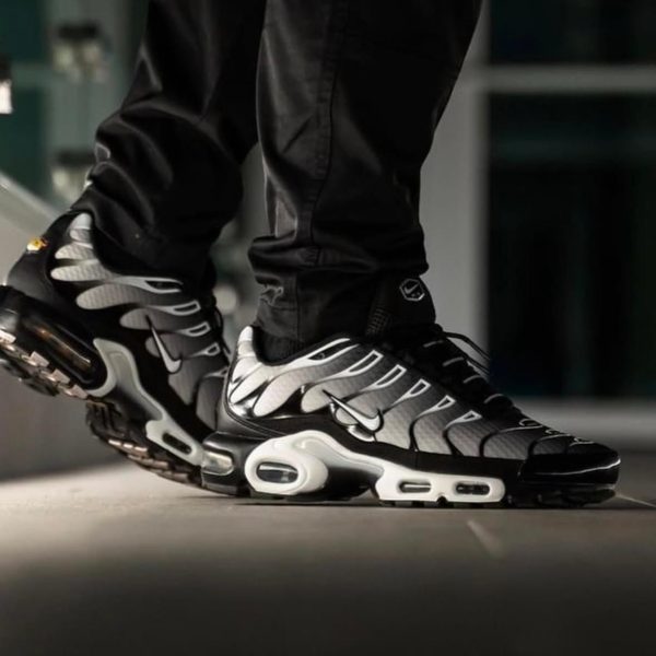 Black and white athletic sneakers with visible air-cushion soles, worn by a person standing indoors on a smooth surface.