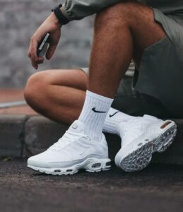 WhatsApp_Image_2025-02-24_at_16.04.41 | UAE SPORT STORE Close-up of a person wearing white Nike sneakers and socks, sitting on steps while holding a phone.