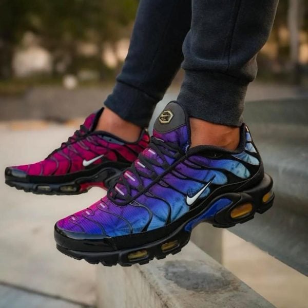 Person wearing colorful gradient Nike sneakers (pink, purple, blue) balancing on a concrete ledge outdoors with dark joggers nearby.