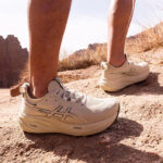 Person wearing light-colored athletic running shoes on rocky desert trail, mid-step on a dusty path with dry grasses in the background.