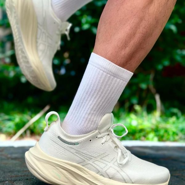 Person wearing a white athletic sneaker with a white sock, captured mid-step on pavement with a green, blurred outdoor background
