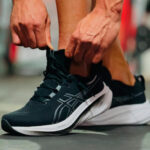 Close-up of hands tying the laces of black running shoes on a gym floor.