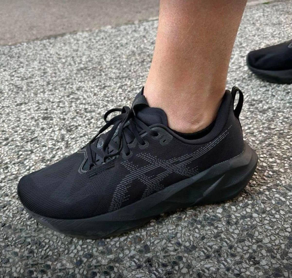 Black athletic sneaker with laces on a person’s ankle, standing on a coarse gravel surface.