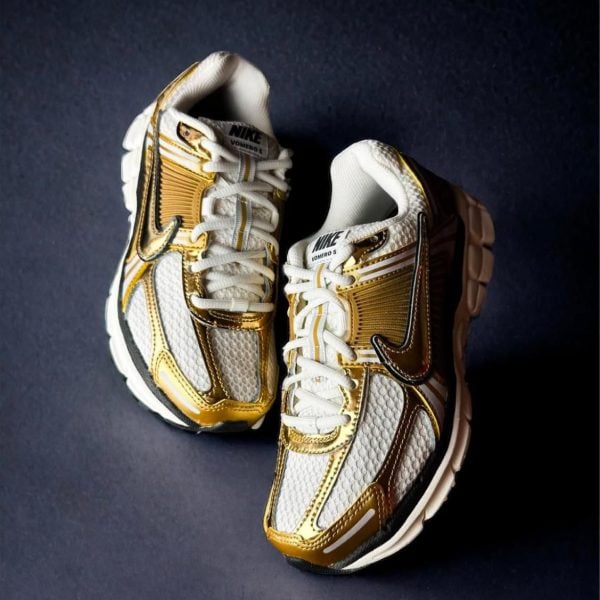 Pair of white and gold Nike VOMERO 5 running shoes on a dark background.