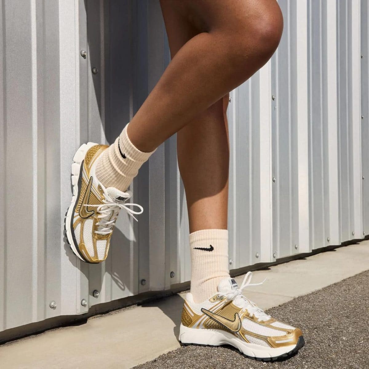 Close-up of a person wearing white and gold Nike sneakers with cream socks, standing against a metal wall.