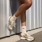 WhatsApp_Image_2025-04-03_at_16.33.06 | UAE SPORT STORE Close-up of a person wearing white and gold Nike sneakers with cream socks, standing against a metal wall.