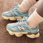 Teal New Balance sneakers with beige accents on a brown carpet, feet crossed.