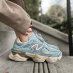 Close-up of a light-blue New Balance sneaker with a chunky beige sole on a person wearing beige jogger pants, standing on concrete steps.