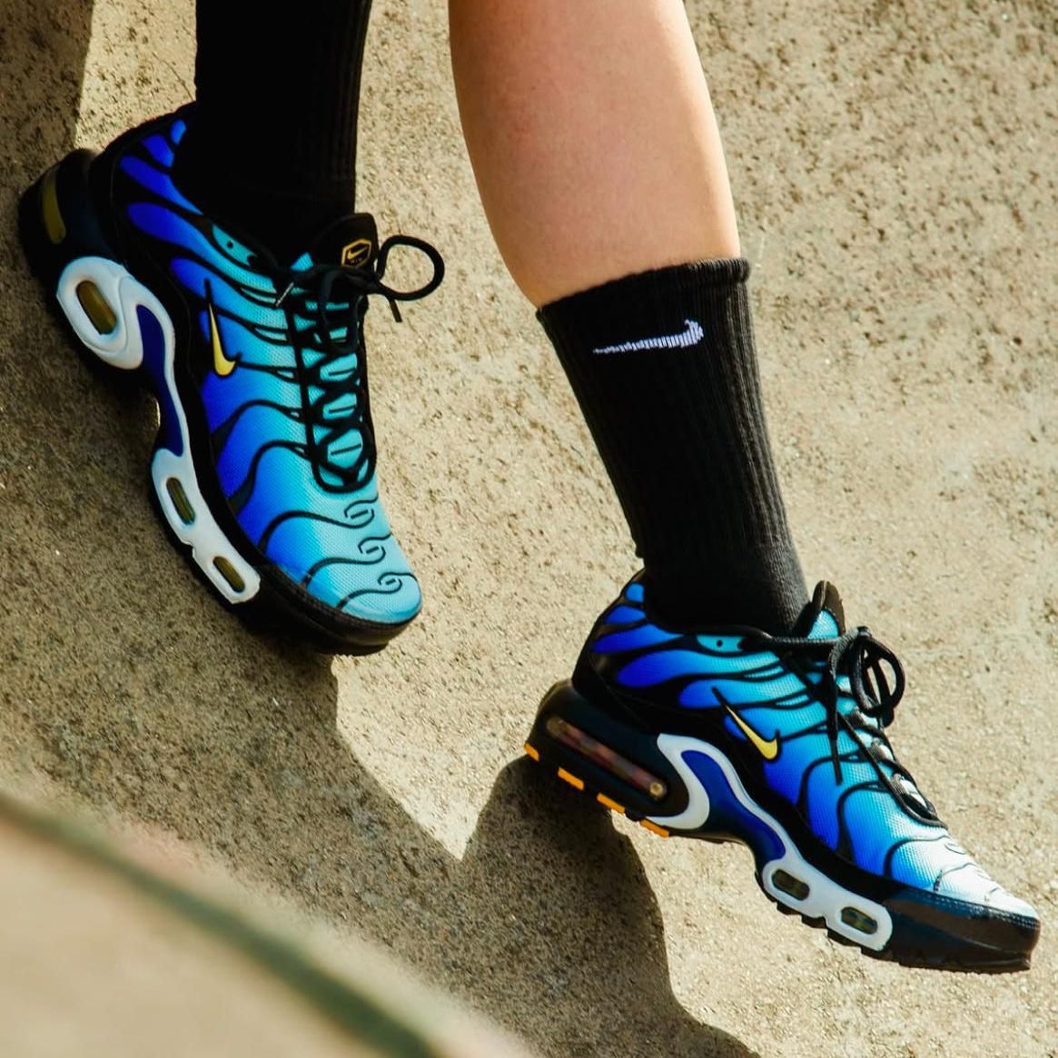 WhatsApp_Image_2025-04-06_at_13.35.02 Pair of blue gradient running shoes with black laces on a sandy surface, mid-step motion showing sole details