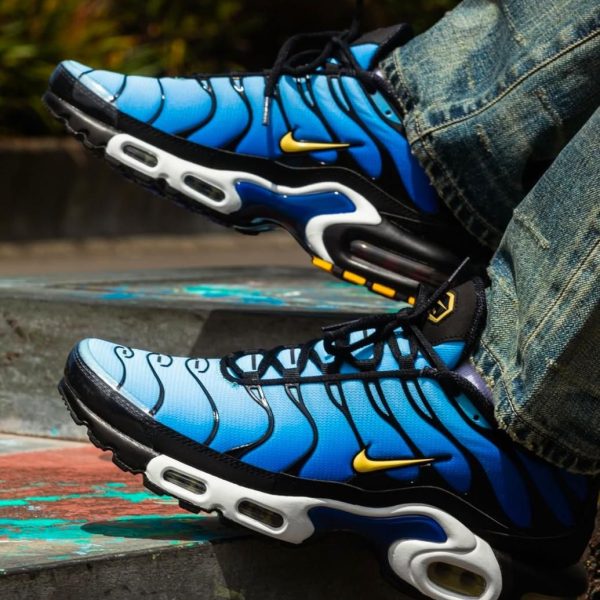 Blue Nike Air Max sneakers with a yellow swoosh resting on a painted concrete step, denim-clad feet in view.