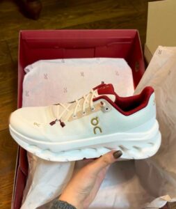 White On running shoe with burgundy lining held above a pink shoebox, showing logo on side.