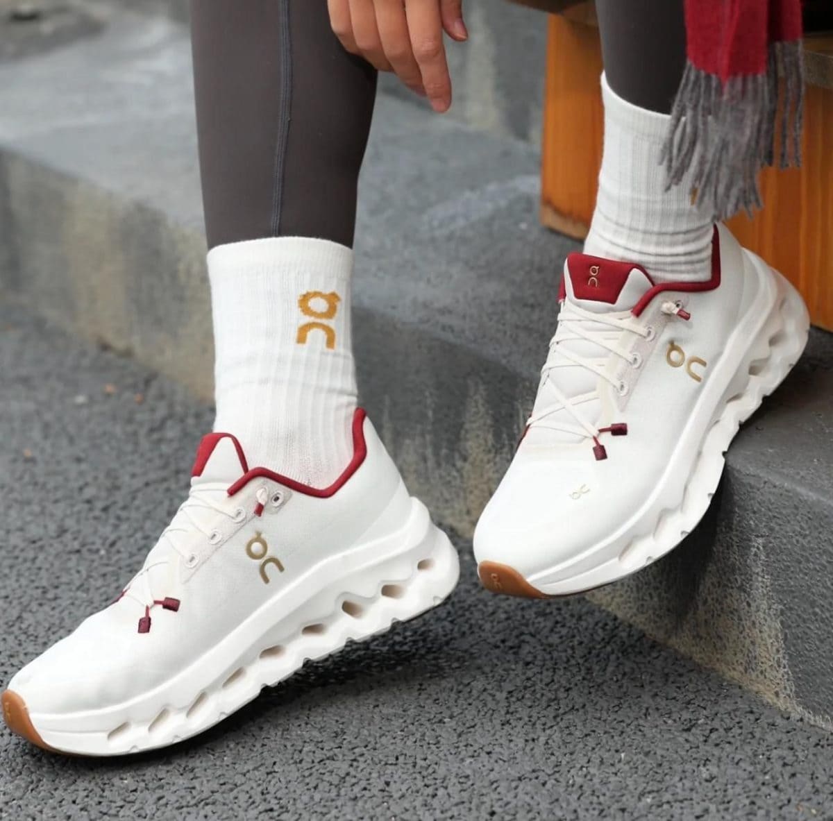 White athletic sneakers with red accents and On logo, worn with white socks and dark leggings outdoors on steps