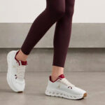 Close-up of a person wearing white running shoes with burgundy accents and burgundy leggings, stepping forward on a light floor.