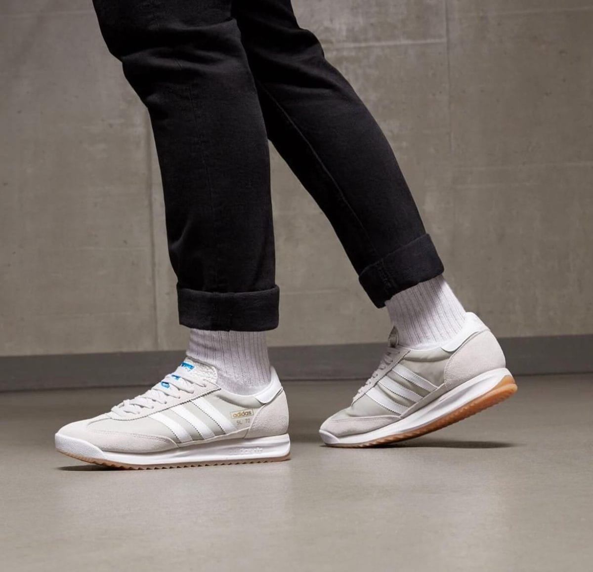 Person wearing white Adidas sneakers with light brown gum soles, gray socks, and black rolled-up jeans standing indoors on a smooth floor.