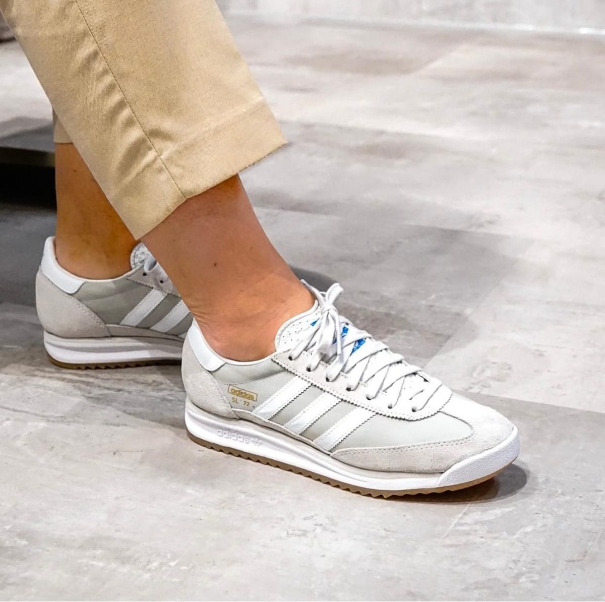 Close-up of a person wearing light gray Adidas sneakers with white stripes and beige cuffed pants, standing on a concrete floor.