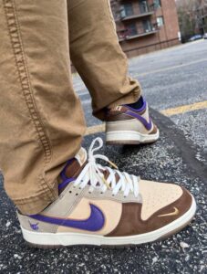Close-up of tan, brown, and purple Nike sneakers with white laces on a person standing on an asphalt road.