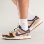 Close-up of beige and brown sneakers with a purple Nike swoosh, worn with white ribbed socks against a neutral background.
