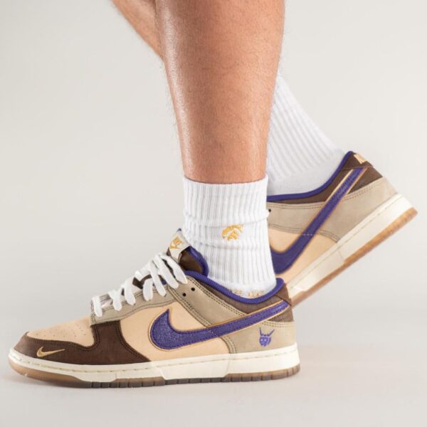 Close-up of beige and brown sneakers with a purple Nike swoosh, worn with white ribbed socks against a neutral background.