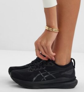 Close-up of a person tying the laces of a black running shoe on their foot, with a white sleeve and gold bracelets visible.