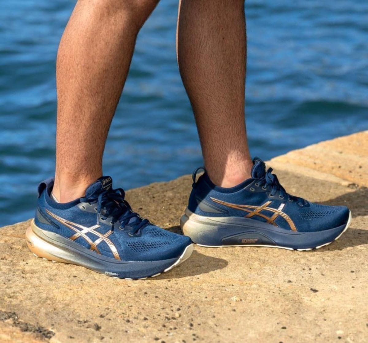 Blue ASICS running shoes on both feet, standing on a rocky shore with water in the background.