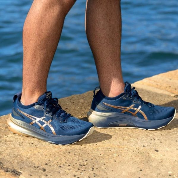Blue ASICS running shoes on both feet, standing on a rocky shore with water in the background.