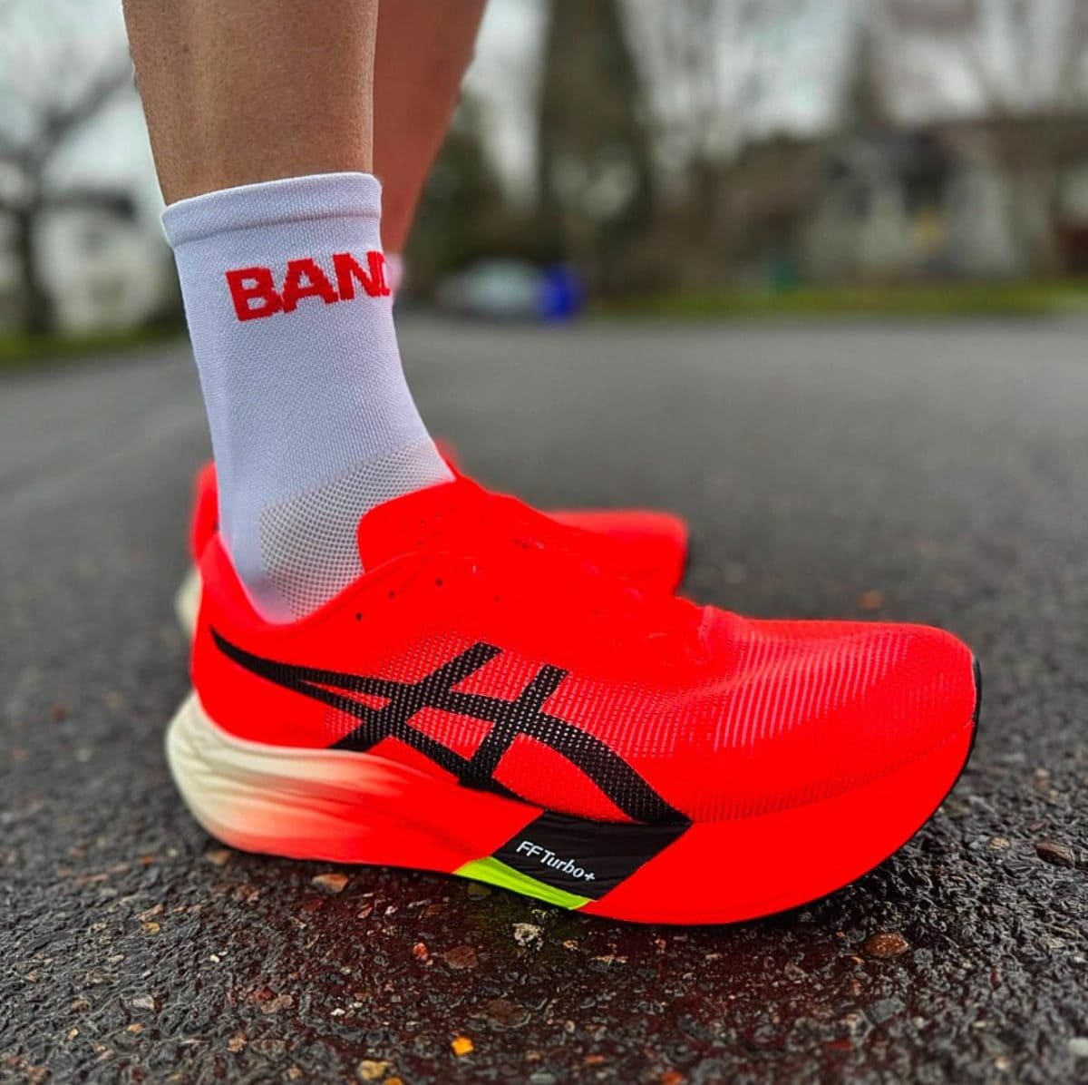 Bright neon red running shoe with black logo on a white sock, standing on a paved road surface.