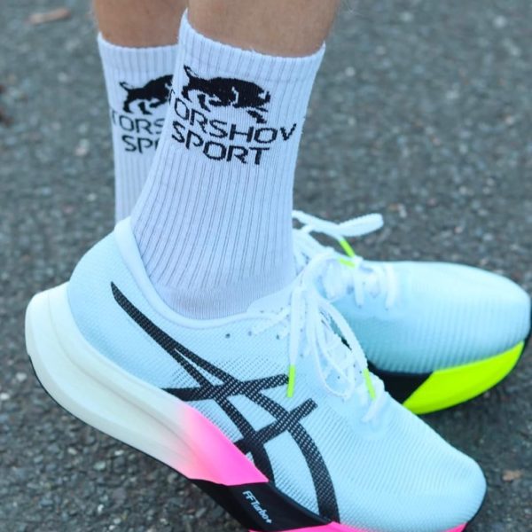 Pair of white running shoes with neon pink outsoles on a dark asphalt surface, worn with white ribbed socks.