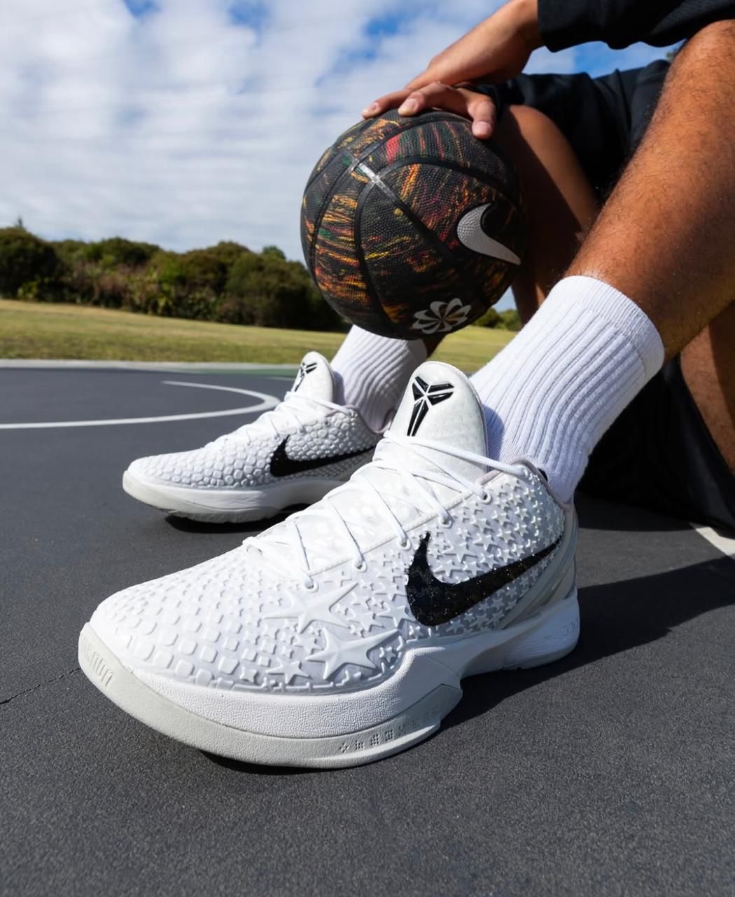 WhatsApp_Image_2025-05-21_at_13.08.20 | UAE SPORT STORE Close-up of white textured Nike basketball shoes with black swoosh, a multicolor basketball, and a player's leg on an outdoor court.