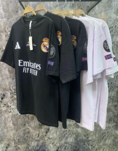 Black Real Madrid T-shirt with Emirates Fly Better slogan on a rack of tees, shown beside white versions, hanging on wooden hangers.