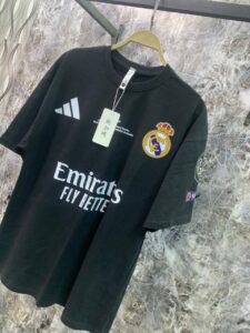 Black Real Madrid football jersey on a hanger featuring Adidas logo and Emirates Fly Better sponsor, with Real Madrid crest on the chest and a price tag attached.