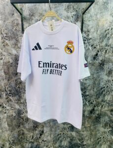 White Real Madrid soccer jersey on a hanger, showing the Emirates sponsor and club crest against a marble background (front view).